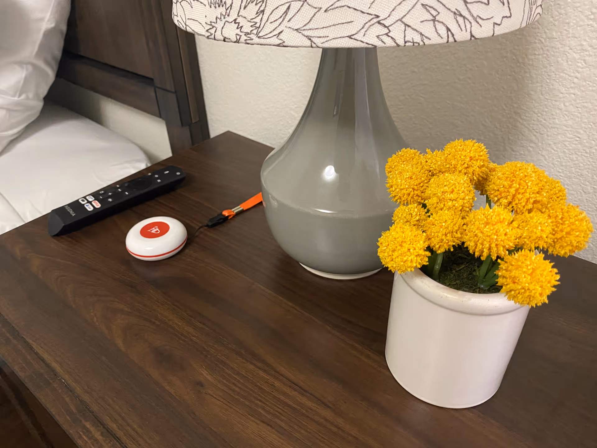 A wooden bedside table with a gray lamp with a floral-patterned lampshade, a small white pot containing bright yellow flowers, a TV remote, and a red and white emergency alert button with an orange lanyard. Part of a bed with white bedding is visible on the left side.