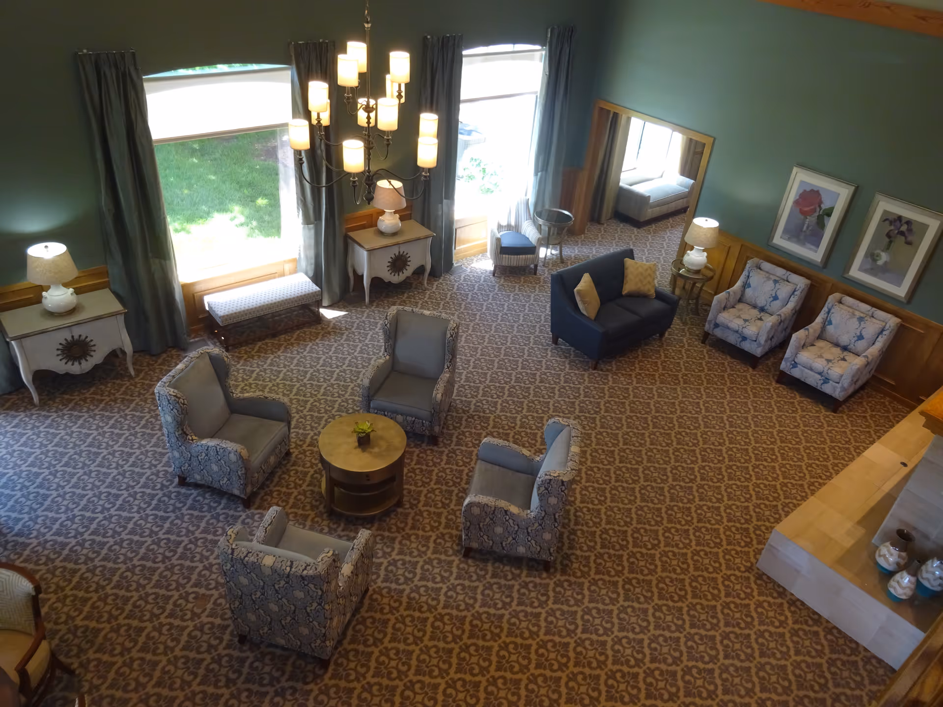 A spacious living room with patterned carpet, several upholstered armchairs arranged around a small round table, a dark blue loveseat with yellow pillows, two side tables with lamps, large windows with curtains letting in natural light, framed floral artwork on the walls, and a chandelier hanging from the ceiling.
