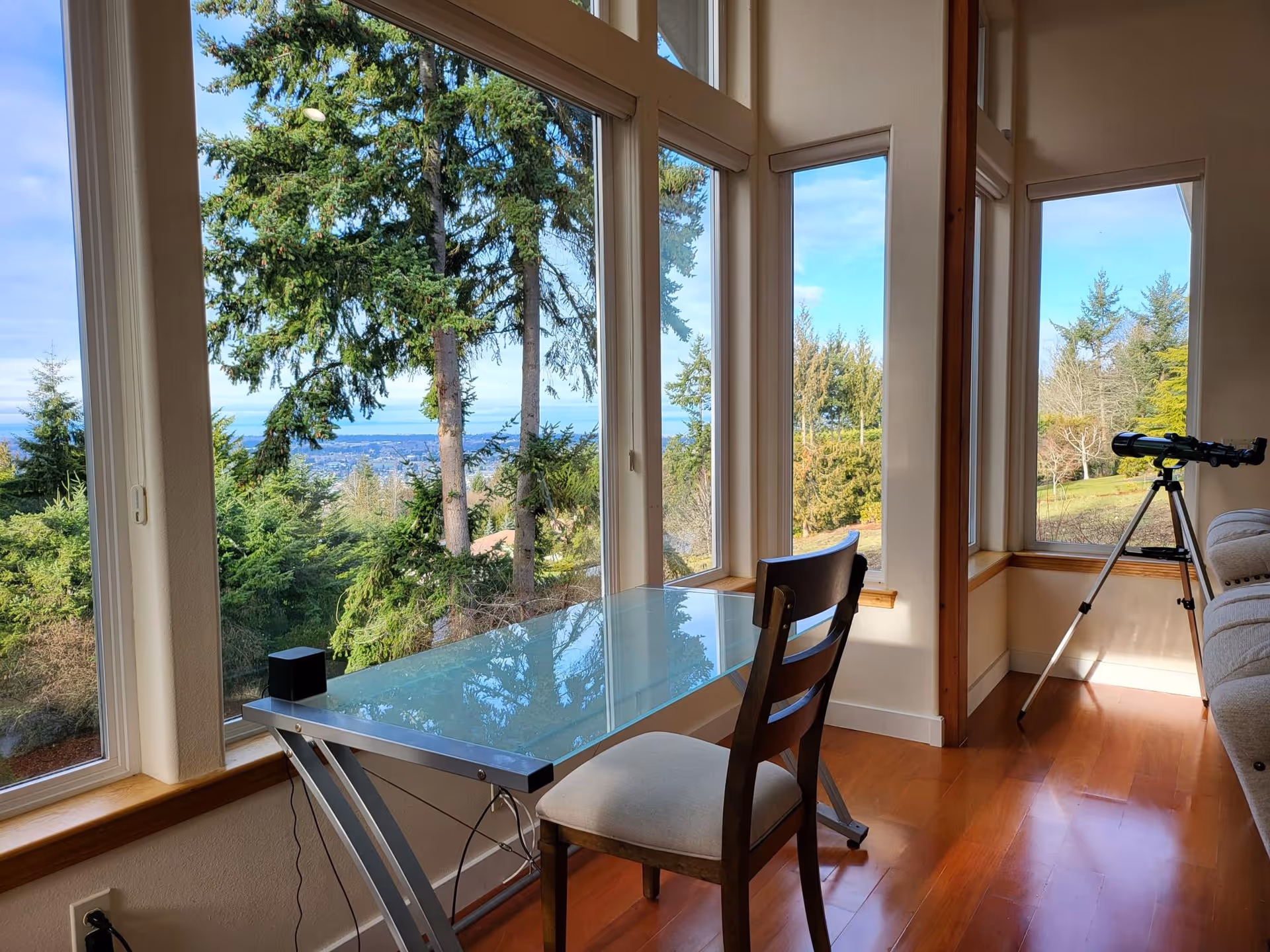 Sunlit interior seating area with a glass desk and chair beside large windows overlooking trees and distant water, and a telescope on a tripod.