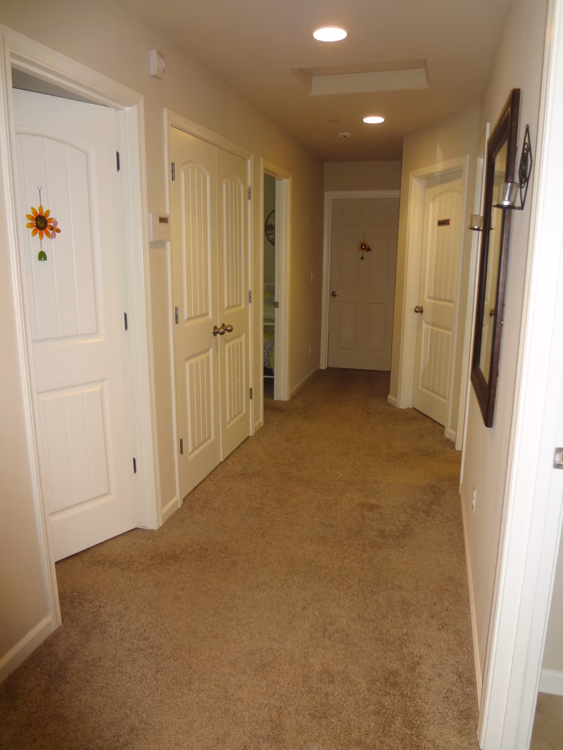 A carpeted hallway in an assisted living facility with multiple closed white doors on both sides. The walls are painted beige, and there is a mirror with candle holders on the right wall. Some doors have small decorative sunflower hangings. The hallway is well-lit with ceiling lights.
