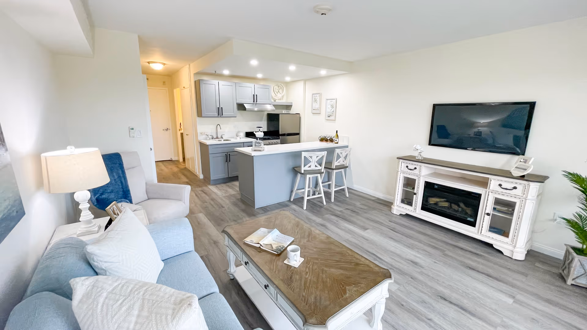 A bright and modern living area in a senior living facility named Ventura Townehouse, featuring a light blue sofa with white pillows, a beige armchair with a blue throw, a wooden coffee table with a book and a cup, a white TV stand with a flat-screen TV mounted on the wall above it, and an open kitchen with gray cabinets, a small island with two bar stools, and stainless steel appliances.