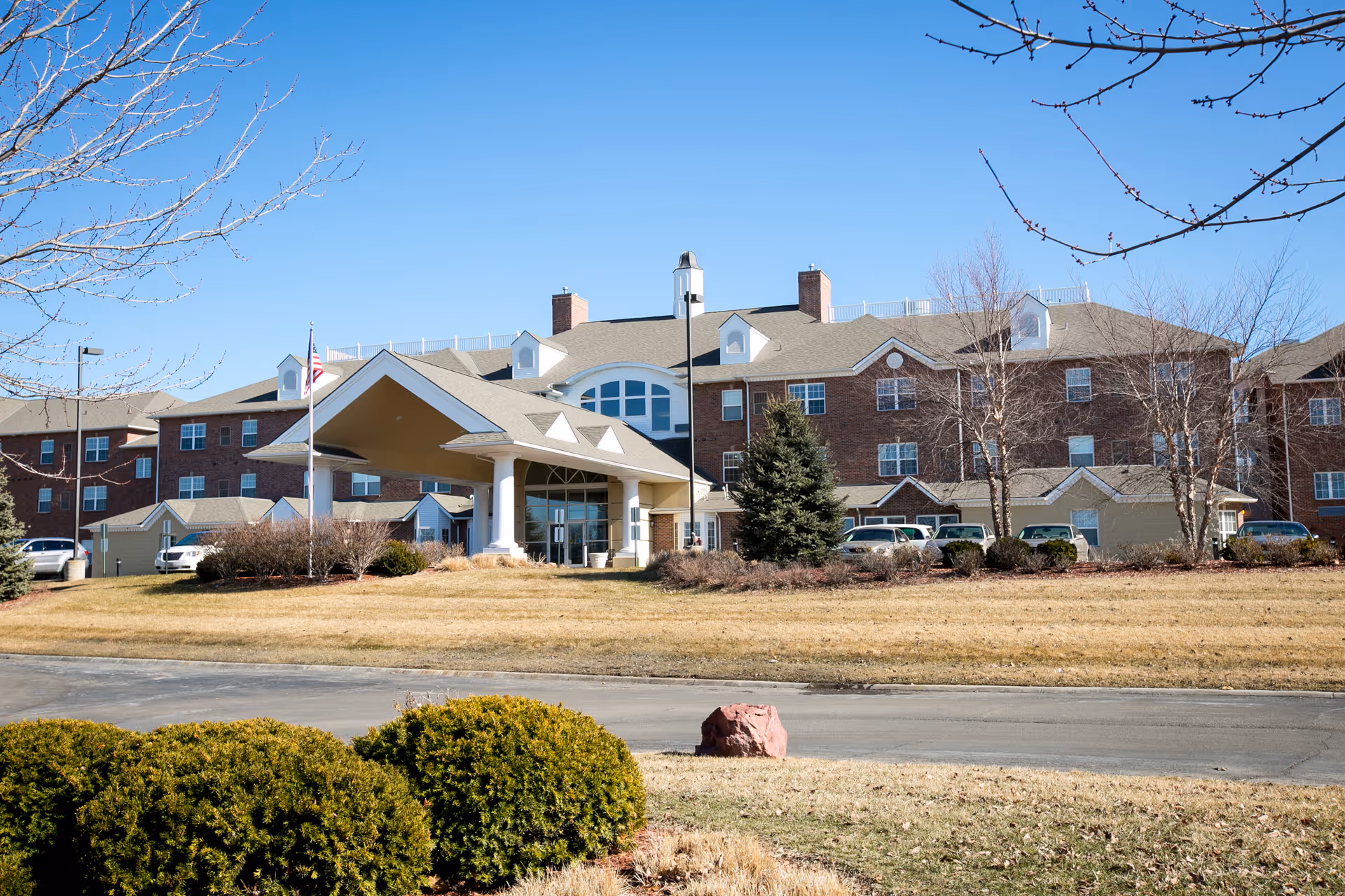 Exterior view of Walnut Grove Retirement Community building with a covered entrance, multiple windows, and a flagpole with an American flag. The landscape includes trimmed bushes, leafless trees, and a clear blue sky.