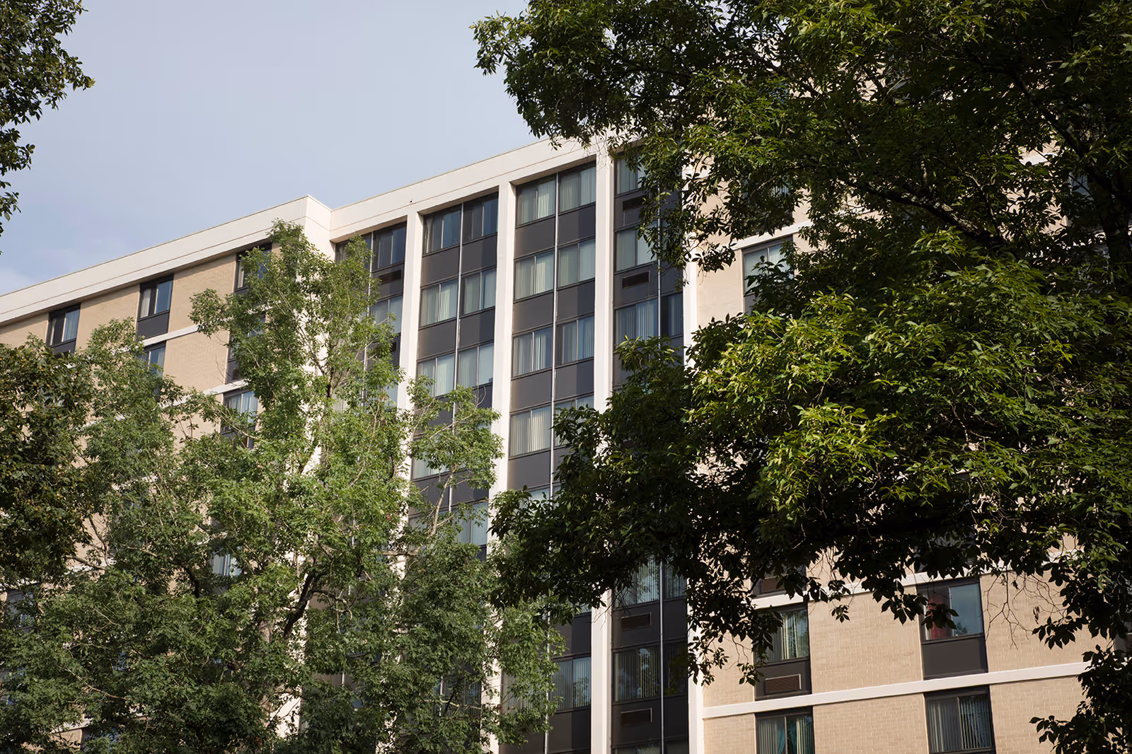 Exterior view of a multi-story senior living facility building partially obscured by green leafy trees under a clear sky.