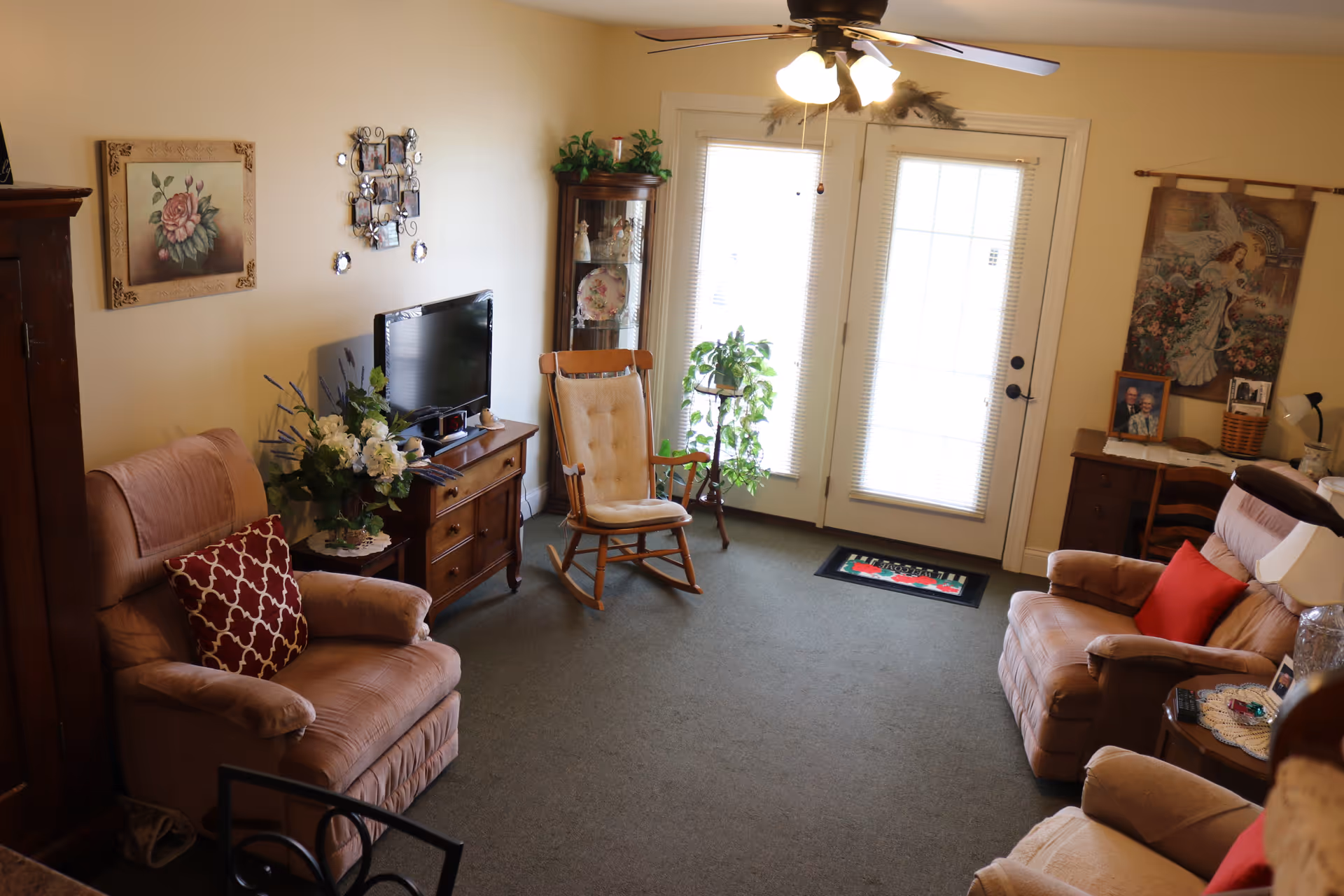 Cozy living room with recliner chairs, a rocking chair, TV on a wooden stand, plants, and French doors letting in light.