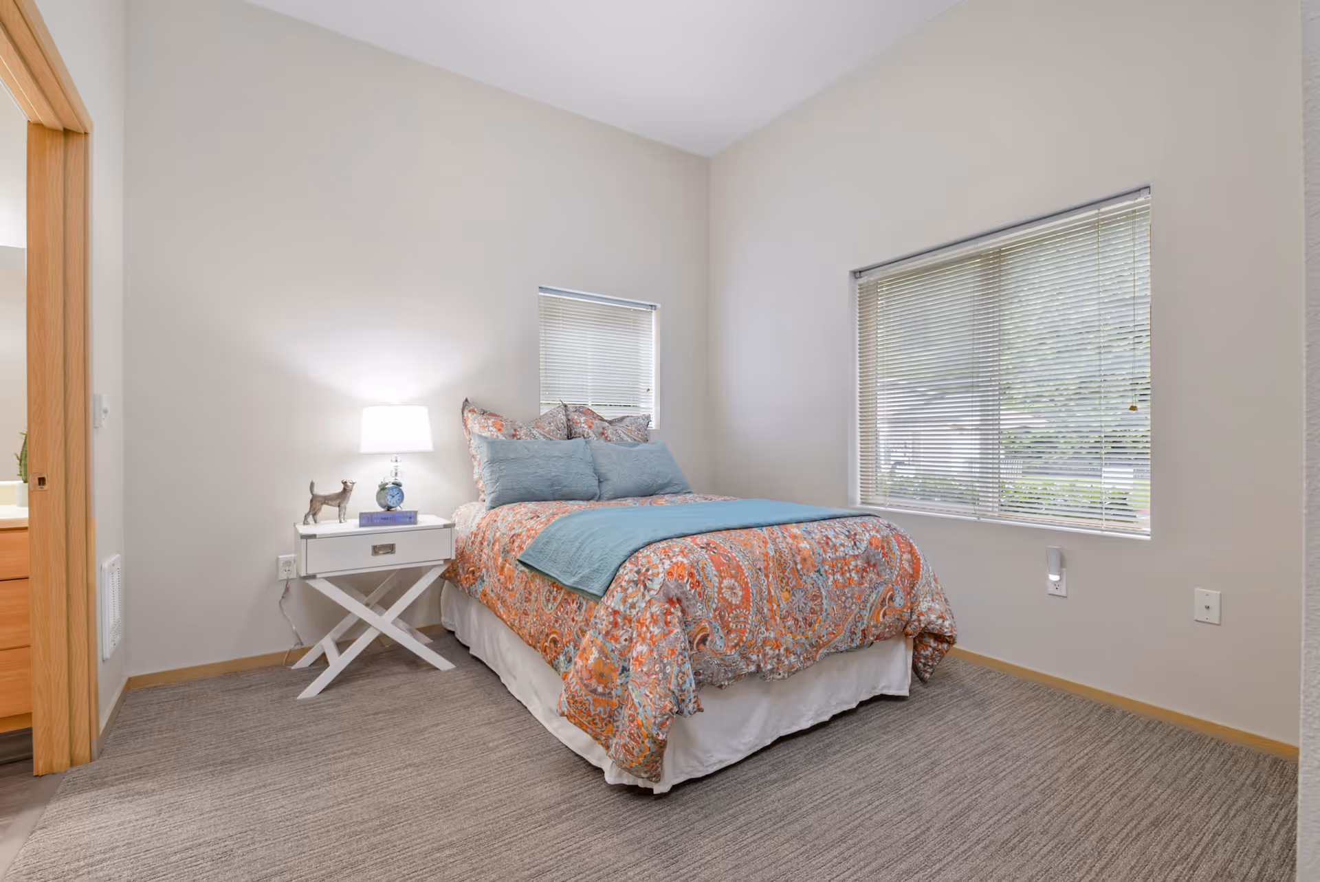 A cozy bedroom with a bed dressed in a colorful patterned comforter and blue pillows. There is a white nightstand with a lamp, a small decorative dog figurine, and a clock. The room has beige walls, a large window with blinds, and a carpeted floor. A doorway leads to an adjacent room.