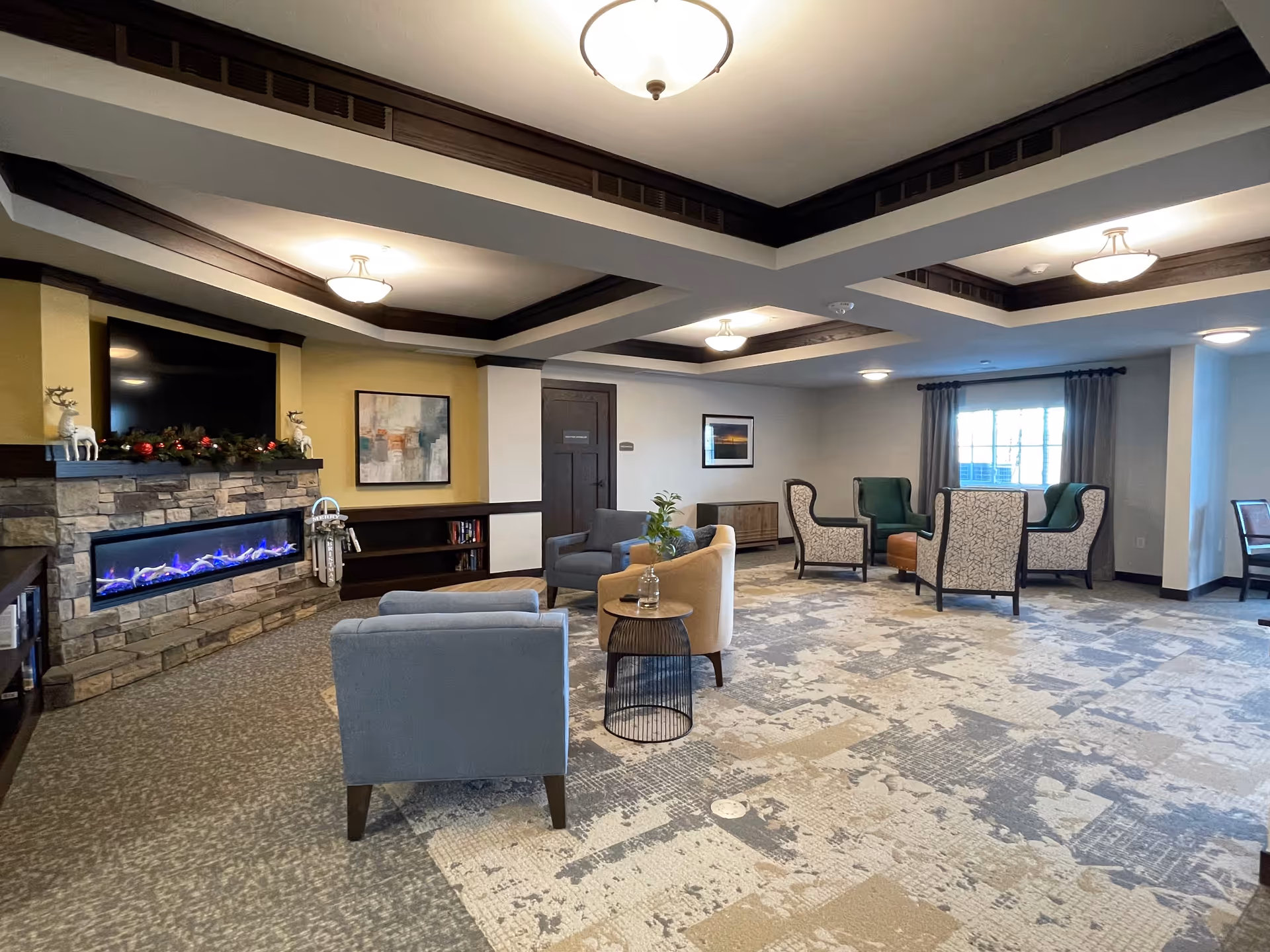 A spacious and well-lit living room area in a senior living facility featuring a modern electric fireplace with stone surround, a mounted flat-screen TV above it, and festive holiday decorations on the mantel. The room has multiple seating arrangements with various upholstered chairs and small tables, patterned carpet flooring, and large windows with curtains allowing natural light. The ceiling has recessed lighting and decorative dark wood trim.