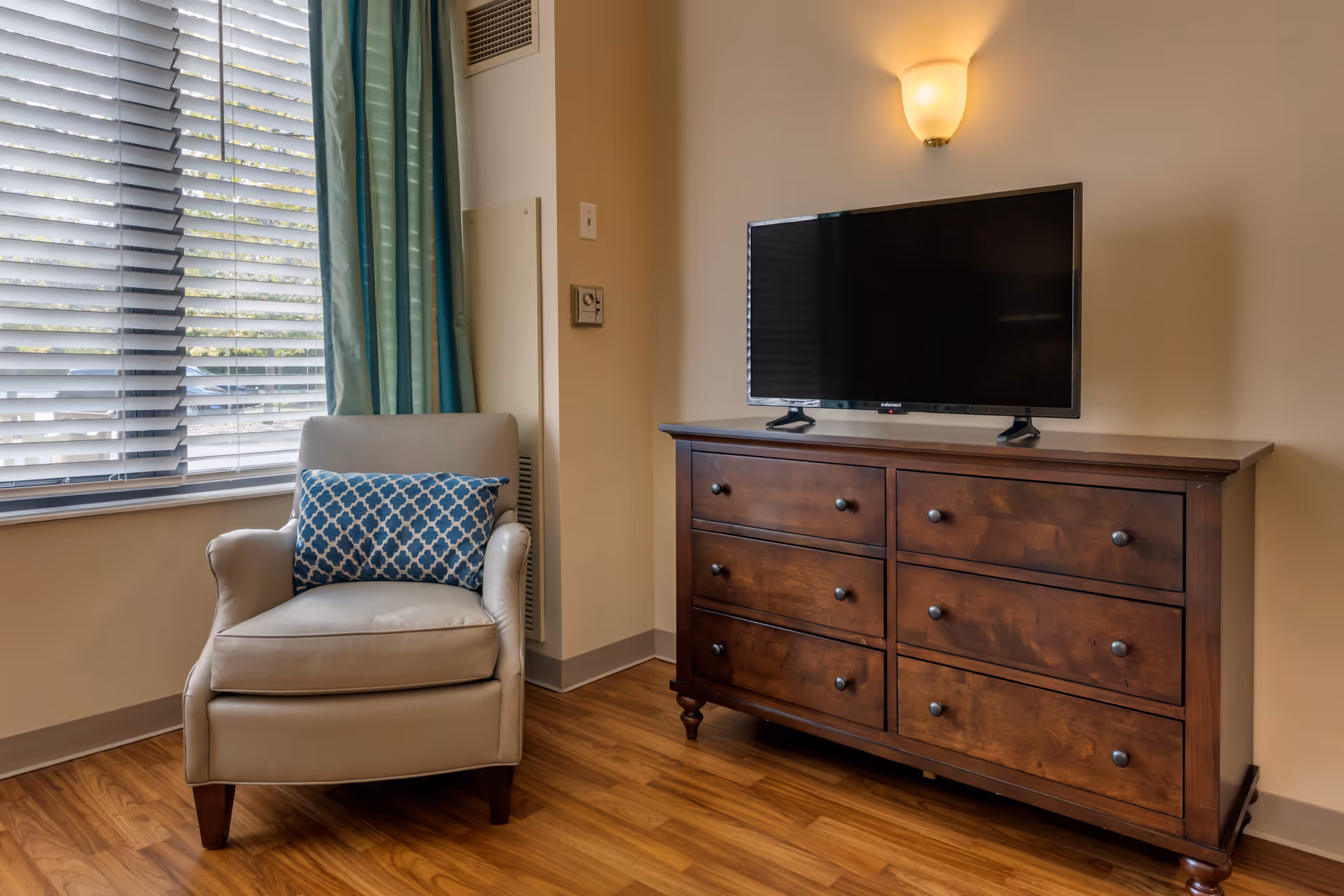 A cozy corner of a room featuring a beige armchair with a blue patterned pillow next to a window with blinds and teal curtains. Adjacent to the chair is a wooden dresser with six drawers, topped with a flat-screen TV mounted on it. A wall sconce light fixture is mounted above the TV, illuminating the beige walls and wood flooring.
