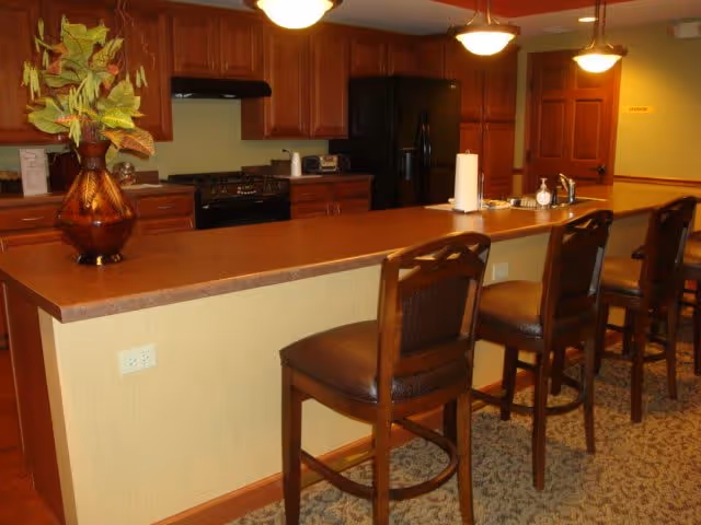 Interior view of a kitchen area with a long counter and four high chairs. The kitchen features wooden cabinets, a black refrigerator, a stove with a hood, and a sink on the counter. There is a decorative plant on the counter and warm lighting from ceiling fixtures.