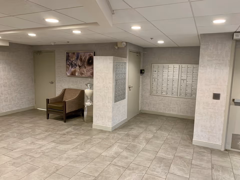Interior lobby/mail area with wall mailboxes, a single armchair, decorative artwork and tiled floor.