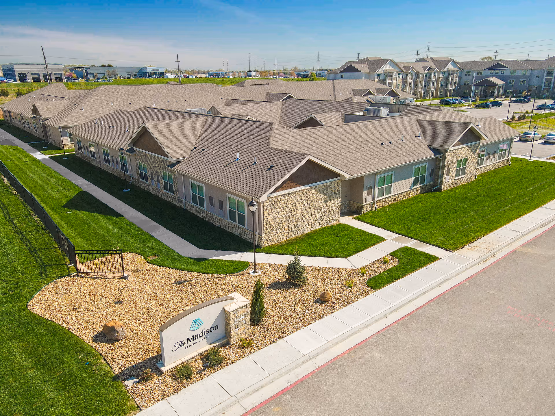 Aerial view of The Madison Senior Living facility showing a large single-story building with a stone and beige exterior, surrounded by well-maintained green lawns, sidewalks, and a parking area in the background. A sign with the facility's name is visible near the entrance.