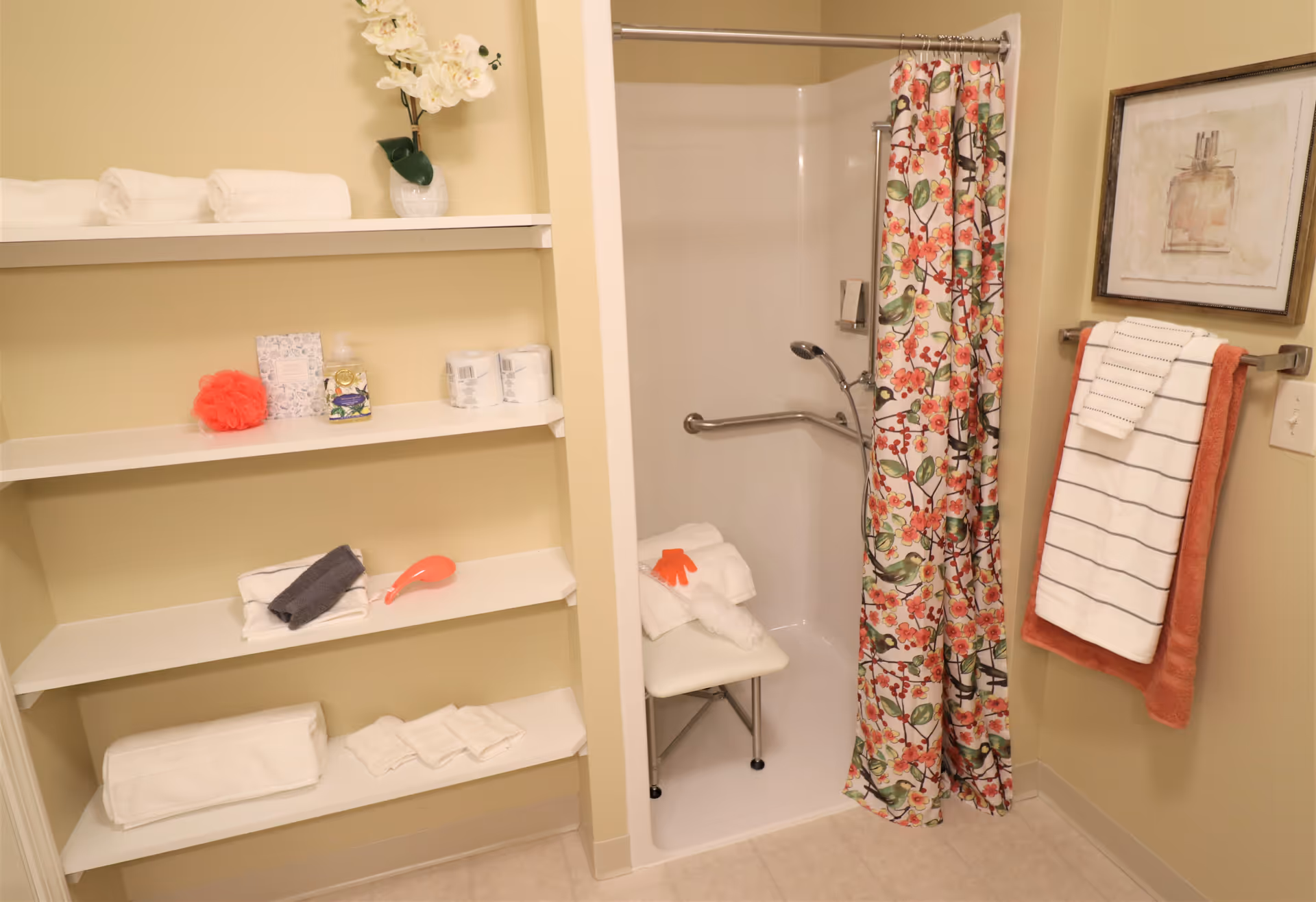 A bathroom with a walk-in shower featuring a floral shower curtain and a white shower chair inside. There are white shelves on the left side holding folded towels, a red bath sponge, toilet paper rolls, and a decorative vase with white flowers. On the right wall, there is a towel rack with two towels hanging and a framed picture above it.