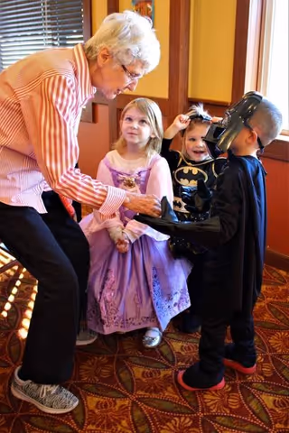 An elderly woman bending down and interacting with three young children dressed in costumes inside a room with patterned carpet and wooden paneling. One child is dressed as a princess in a purple dress, another as Batman, and the third as Darth Vader.