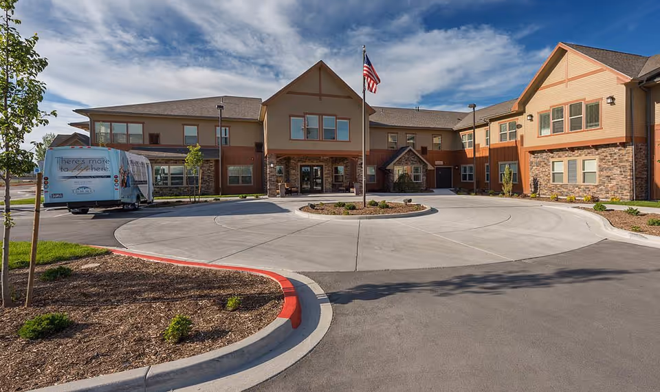 Exterior view of a senior living facility building with a circular driveway and an American flagpole in the center. The building has two stories with a mix of stone and beige siding, and there is a shuttle bus parked on the left side. The sky is partly cloudy with blue patches.