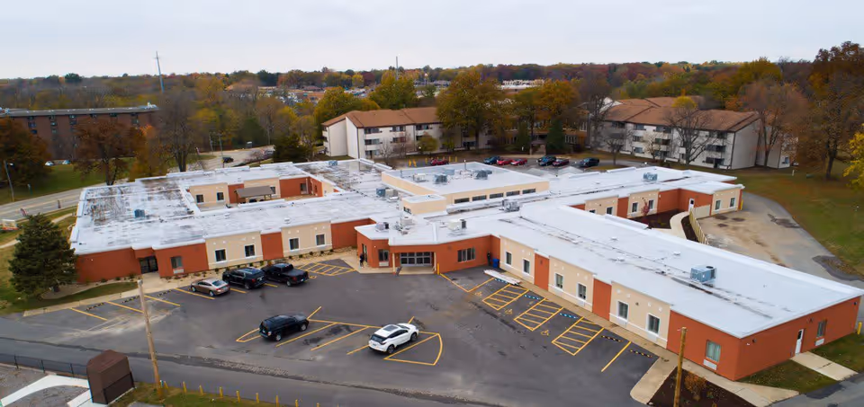 Aerial view of The Loft Rehabilitation of Rock Springs facility showing a large single-story building with a white roof and red and beige exterior walls. The building is surrounded by a parking lot with several cars parked and some trees with autumn foliage in the background.