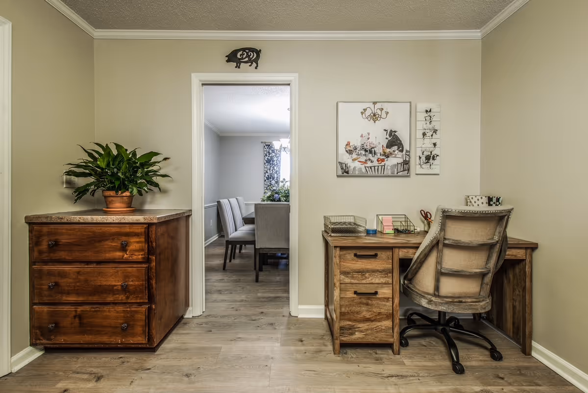 A cozy interior space featuring a wooden desk with an office chair, a wooden chest of drawers with a potted plant on top, and a doorway leading to a dining room with a table and chairs. The walls are beige, and there are two pieces of artwork depicting farm animals above the desk.