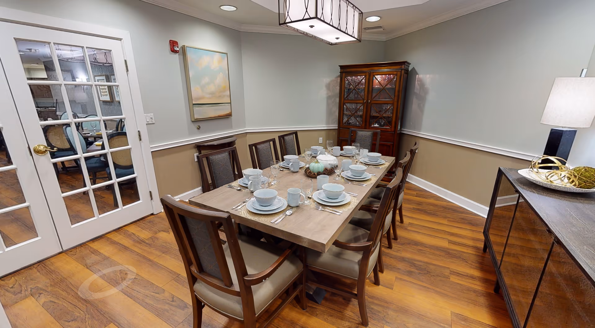 A dining room with a wooden table set for eight people with white dishes, bowls, cups, and silverware. The room has wooden flooring, a glass-front cabinet in the corner, a sideboard with decorative items and a lamp, and a painting on the wall. French doors with glass panes lead to another room with additional seating.
