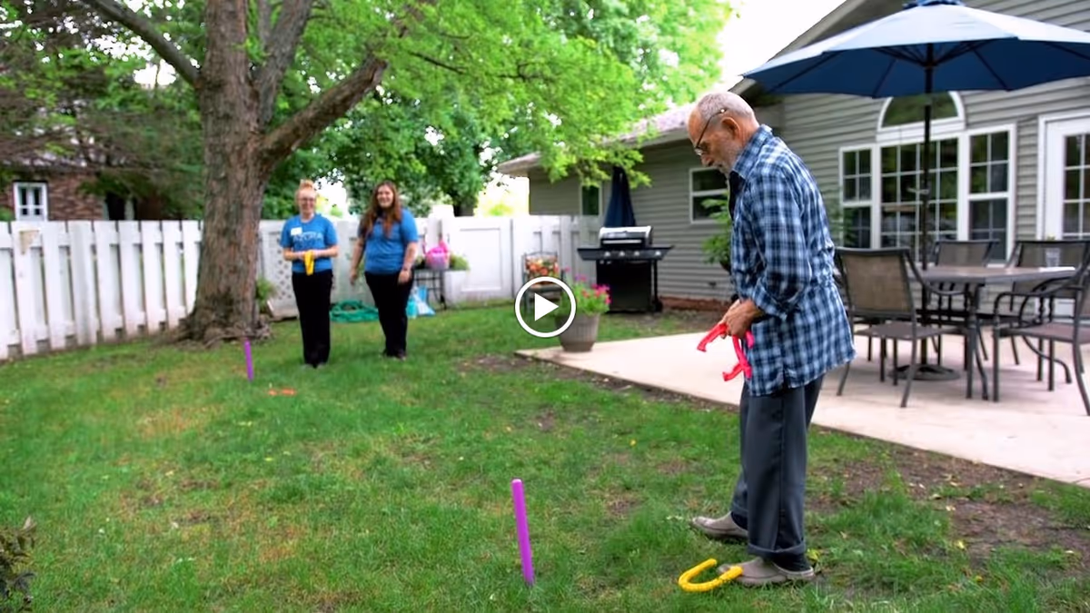 An elderly man playing a ring toss game on a grassy lawn in the backyard of a residential facility. Two women stand in the background near a white fence, watching and smiling. The yard has a large tree, patio furniture with an umbrella, and a barbecue grill near the building.