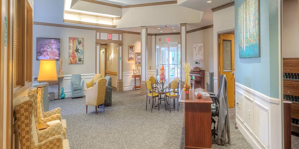 A bright and spacious senior living community common area with comfortable armchairs, a small table with chairs, a reception desk, decorative artwork on the walls, and a television mounted on the wall. The space has carpeted floors, soft lighting, and large windows allowing natural light to enter.