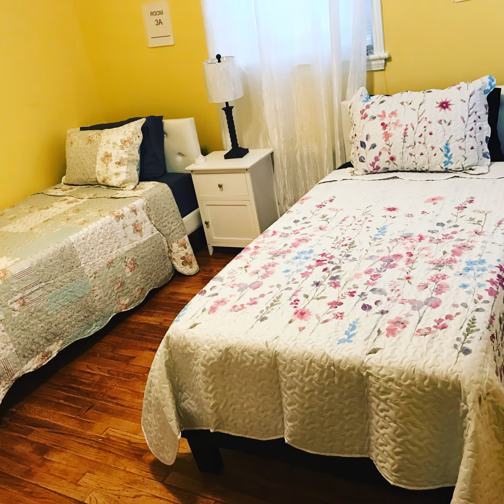 A small bedroom with two single beds separated by a white nightstand with a lamp. One bed has a floral quilt with pink, purple, and blue flowers, and the other has a patchwork quilt in muted tones. The walls are painted yellow, and there is a window with sheer white curtains. A sign on the wall reads 'ROOM 3A'.