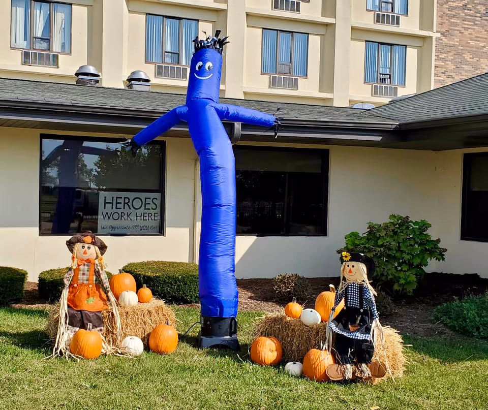 Outdoor scene at a senior living facility with a blue inflatable tube man waving, two scarecrow decorations sitting on hay bales, and several pumpkins arranged on the grass. The building in the background has windows and a sign that reads 'HEROES WORK HERE We appreciate all you do!'