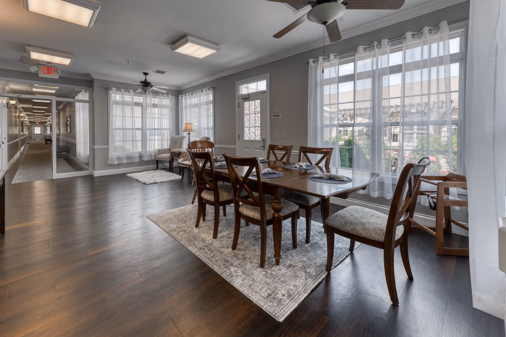 A bright dining area in Addington Place of Shoal Creek featuring a wooden dining table with six chairs set on a patterned rug. Large windows with sheer white curtains allow natural light to fill the room. In the background, there is a hallway with carpeted flooring, a ceiling fan, and a small seating area with two upholstered chairs and a lamp.