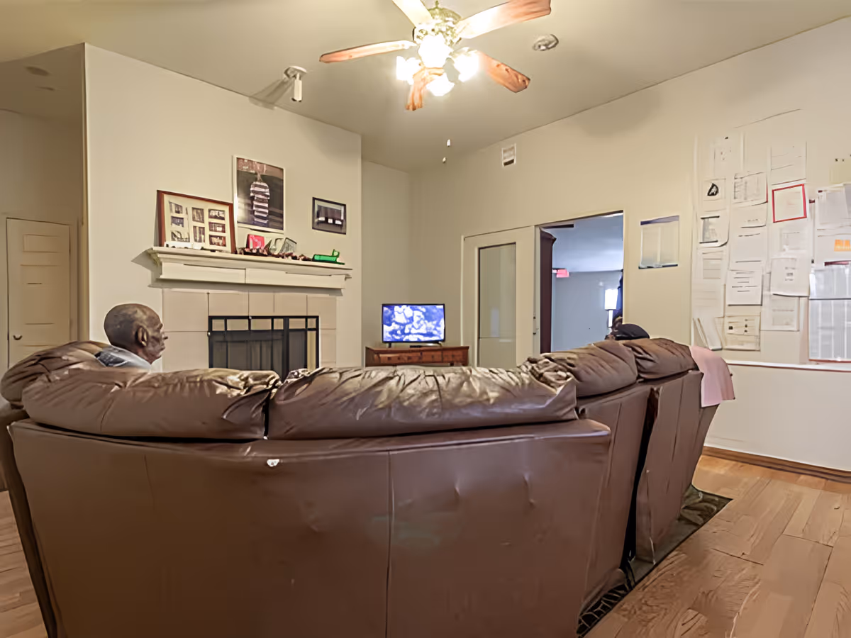 A living room with a brown leather sectional sofa facing a small TV on a wooden stand. There is a fireplace with a white mantel decorated with framed photos and artwork above it. A ceiling fan with lights is on the ceiling. The walls have various papers and documents pinned on one side. Two people are sitting on the sofa, watching the TV.