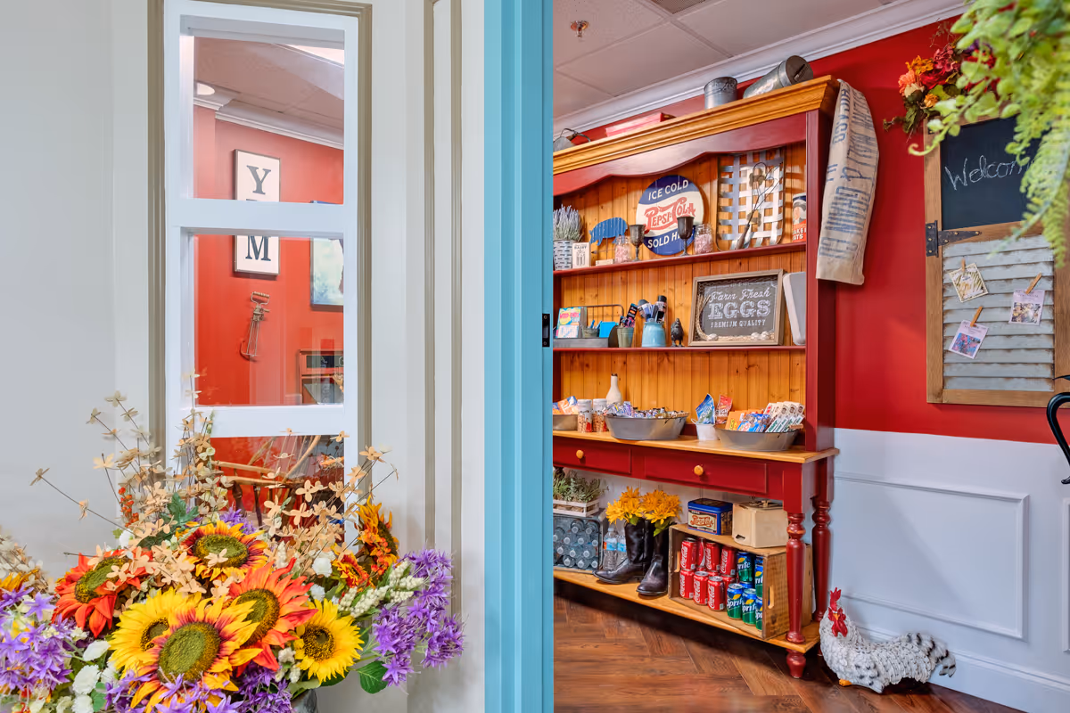 A cozy interior corner featuring a wooden shelf filled with various decorative items, snacks, and drinks. The shelf is painted red with natural wood accents and is set against a red wall. To the left, there is a window with white framing and a colorful bouquet of flowers in front of it. A chalkboard with pinned photos and a welcome message is visible on the wall to the right.
