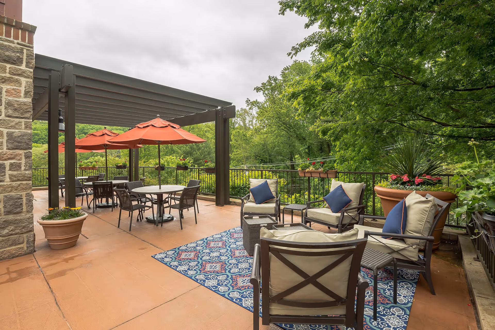 Outdoor patio area with cushioned chairs and small tables on a patterned blue rug, several round tables with orange umbrellas, surrounded by greenery and potted plants under a cloudy sky.