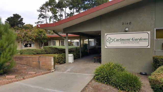 Entrance to Carlmont Gardens Nursing Center showing a covered walkway with a bench, surrounded by bushes and trees, with a sign on the building wall displaying the facility name and address number 2140.