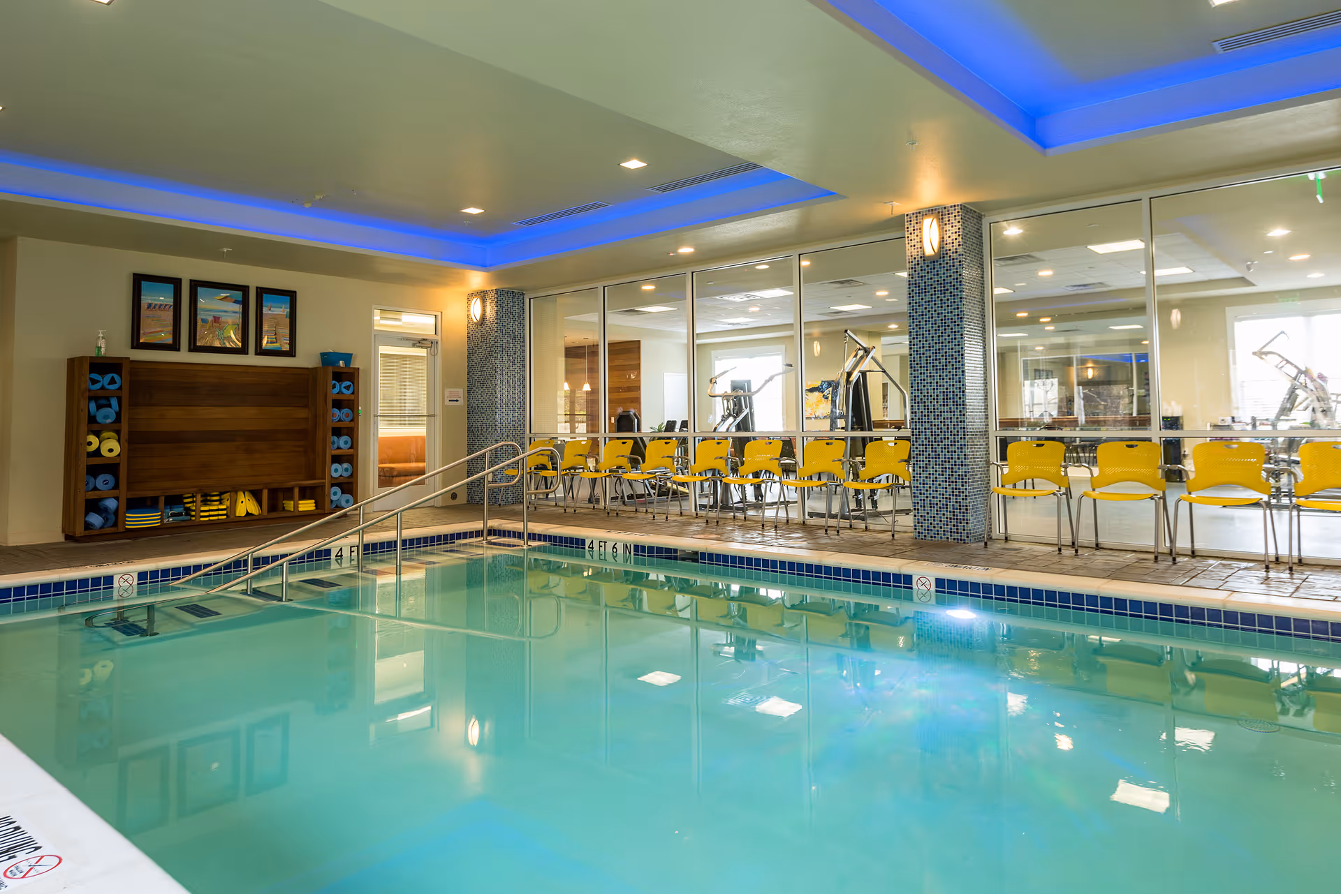 Indoor swimming pool with a metal handrail, yellow chairs along a glass wall and a fitness room visible beyond.