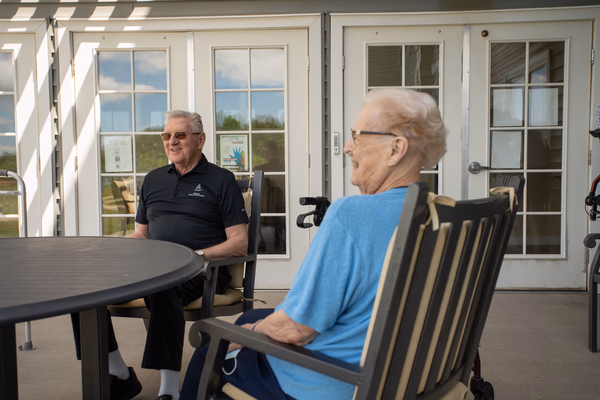 Two elderly individuals sitting and conversing on a covered outdoor patio area with glass-paneled doors in the background. One person is wearing a black polo shirt and sunglasses, and the other is wearing a blue shirt and glasses.