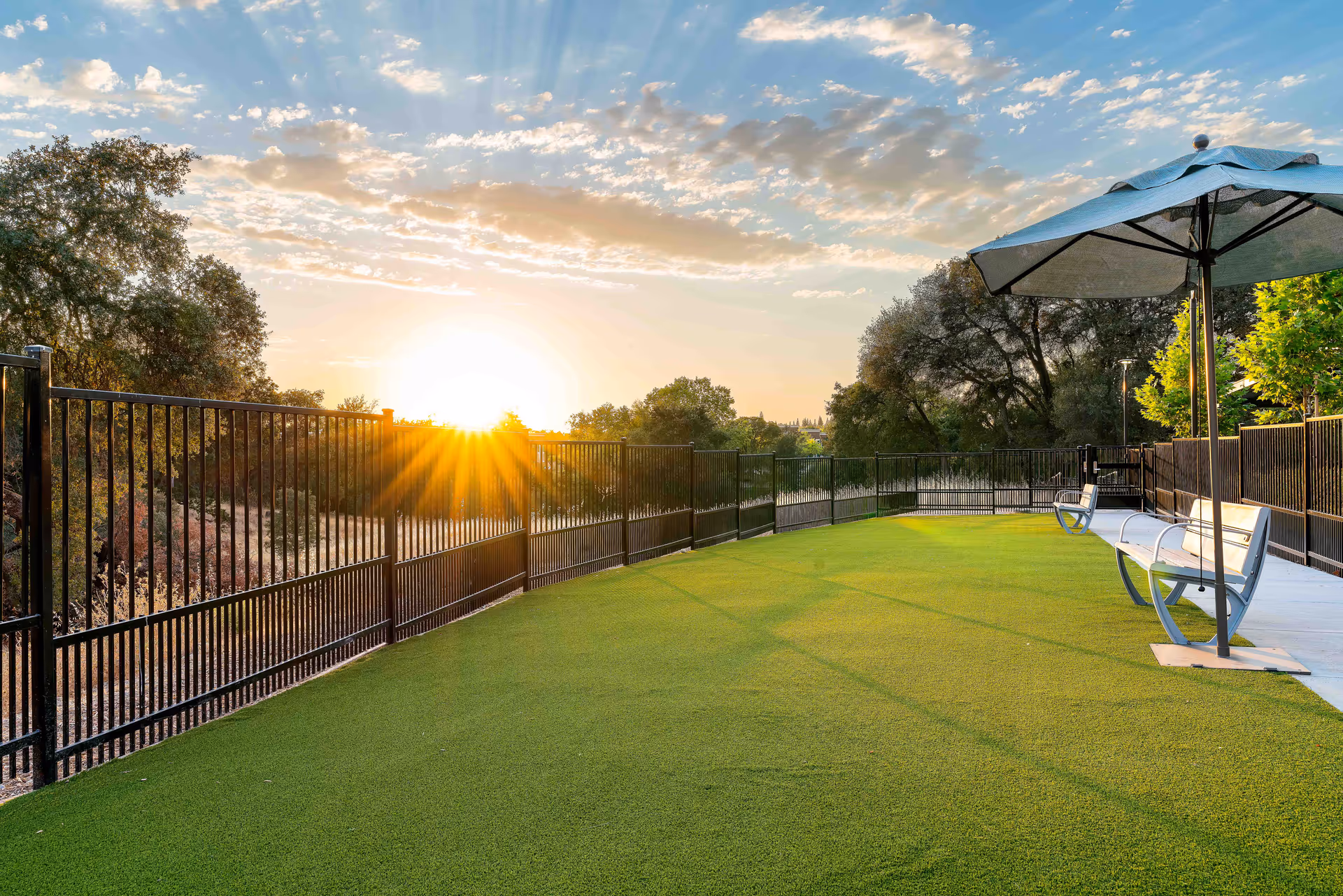 Outdoor fenced area with green artificial turf, two white benches, and a large umbrella on a concrete path, with trees and a sunset sky in the background.