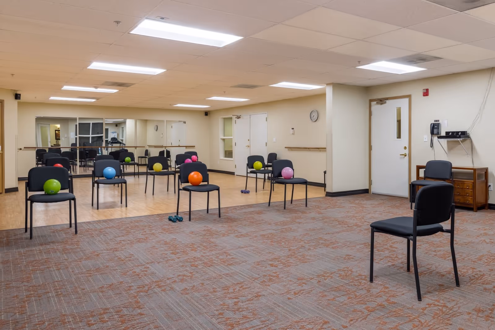 A spacious exercise room with multiple black chairs arranged in rows, each chair holding a colorful exercise ball. The room has a large mirror on one wall, wooden flooring in the exercise area, and carpeted flooring in the foreground. There are also a few small dumbbells on the floor, fluorescent ceiling lights, a clock on the wall, and a door with a phone mounted nearby.