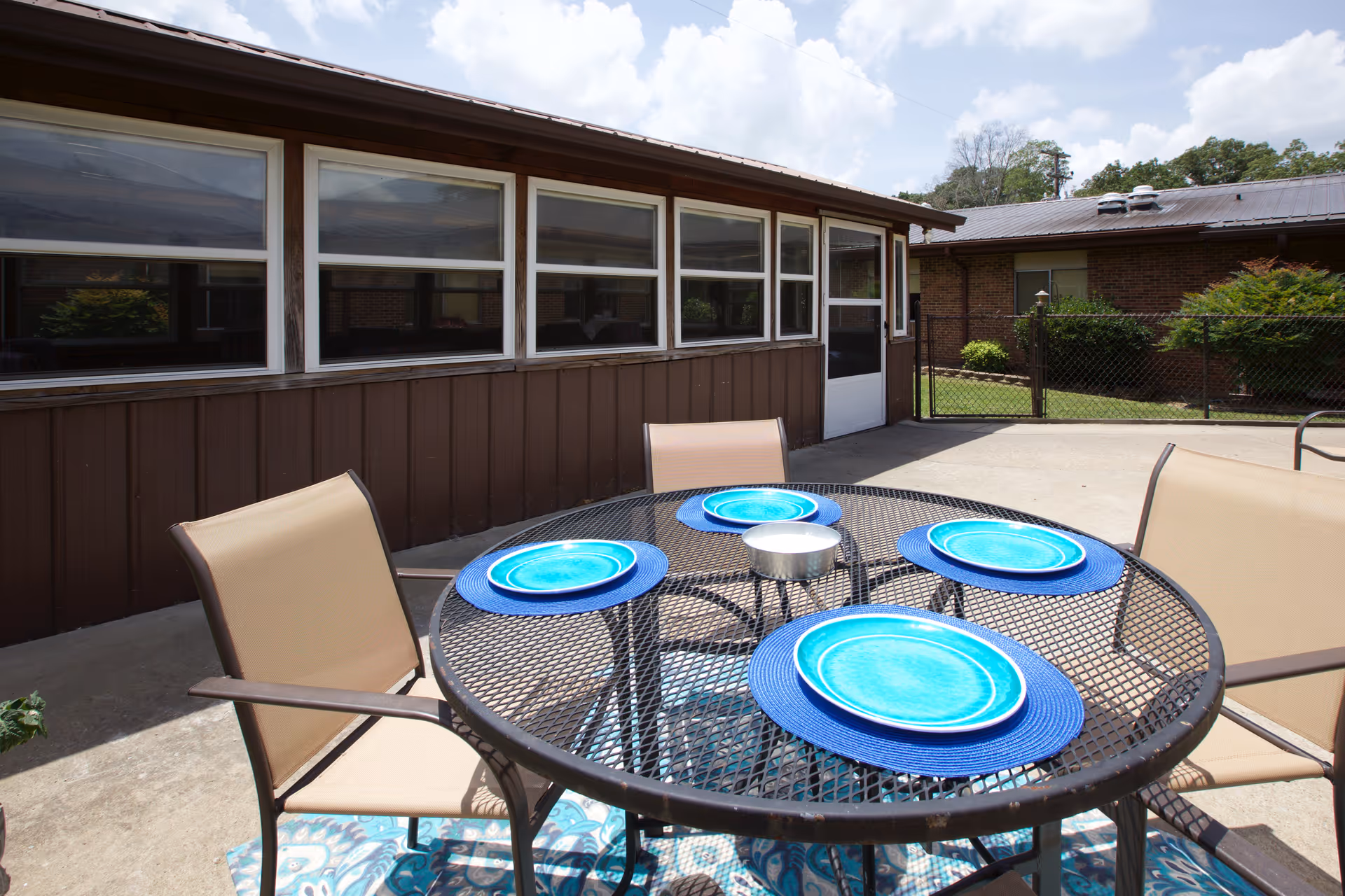 Outdoor patio area with a round metal table set with four blue plates on blue placemats and four beige mesh chairs. The patio is adjacent to a brown building with multiple windows and a white door. There is a chain-link fence and greenery in the background under a partly cloudy sky.