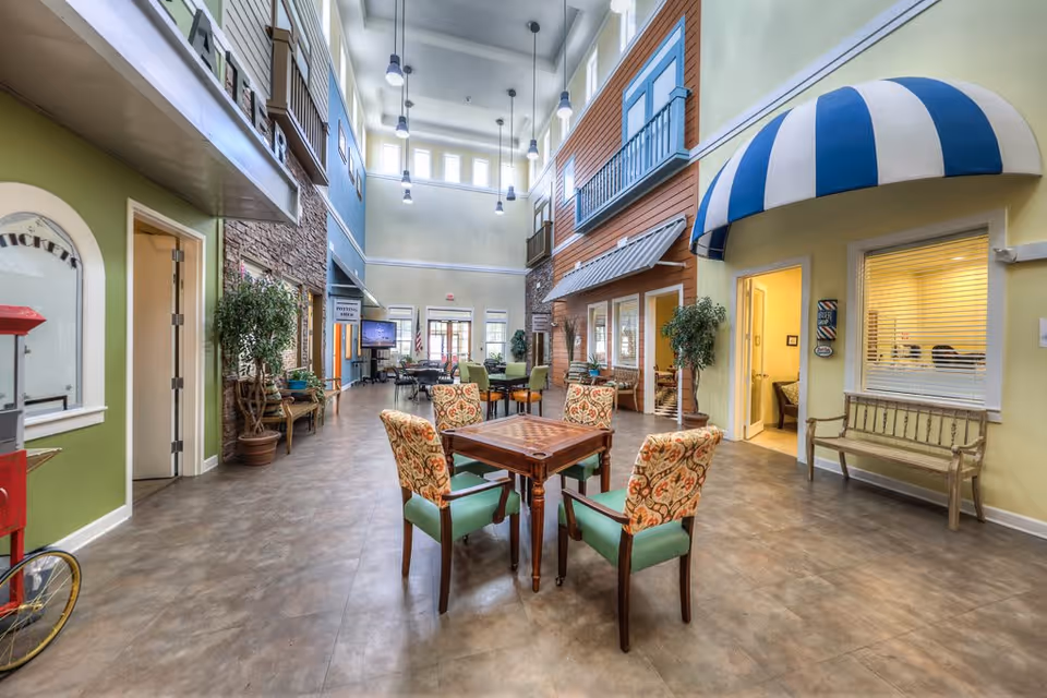 A spacious, well-lit common area in a senior living facility with high ceilings and large windows. The room features a central wooden table with four cushioned chairs, patterned with floral designs. The walls are decorated to resemble an outdoor street scene with faux building facades, balconies, and awnings. There are potted plants and benches along the walls, and a popcorn machine is visible on the left side. The area has a warm and inviting atmosphere.