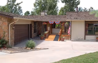 Exterior view of a single-story residential building with a stone and wood facade, a garage on the left, a wooden ramp leading to the entrance decorated with hanging flower pots and patriotic bunting, and an American flag displayed near the entrance.