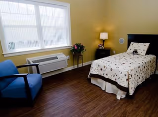 A small bedroom with a single bed covered in a patterned quilt, a blue armchair, a window with vertical blinds, a nightstand with a lamp and flowers, and a wall-mounted air conditioning unit.