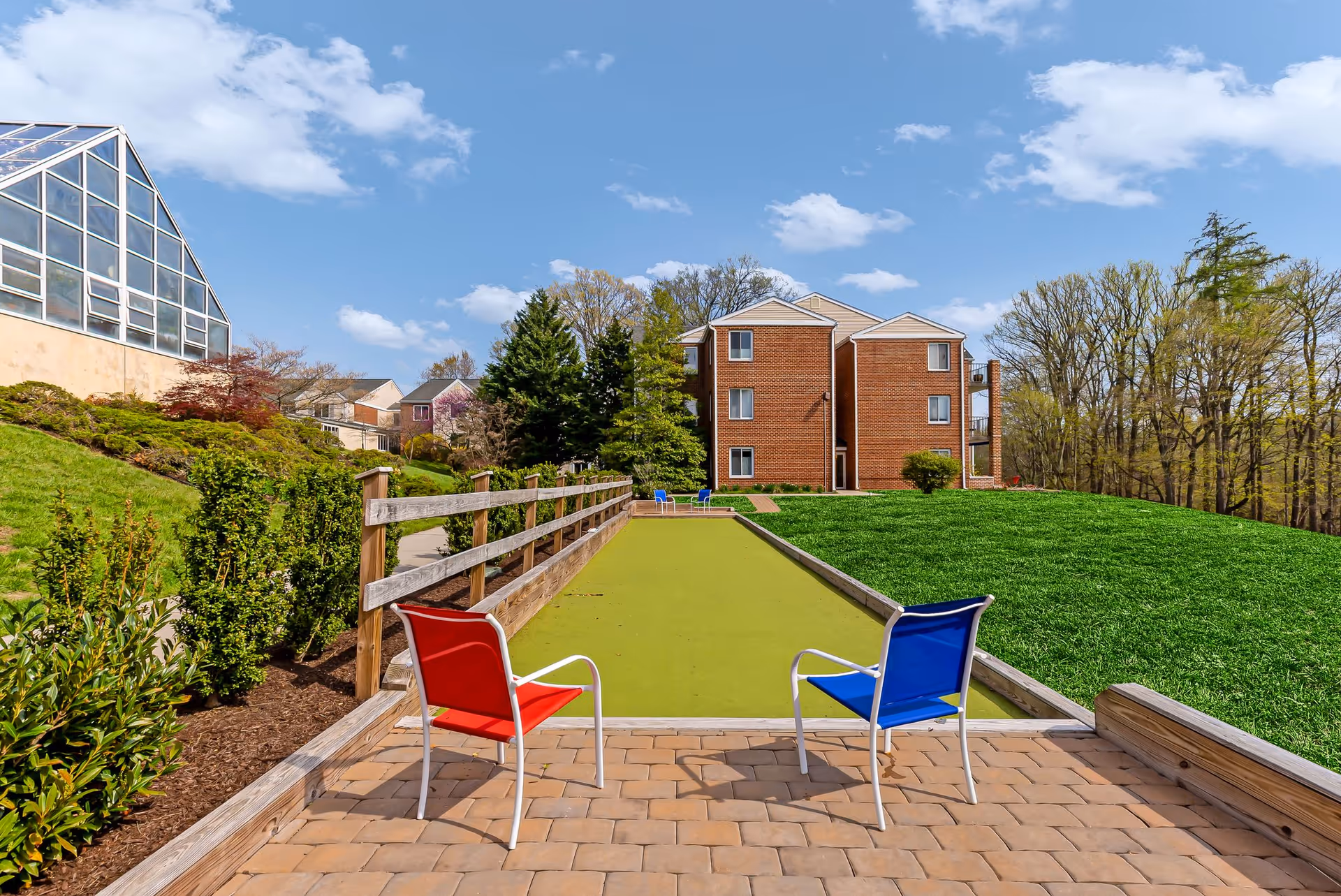 Two colorful lawn chairs face a long bocce court on a paved patio with a brick residential building and landscaped grounds under a blue sky.