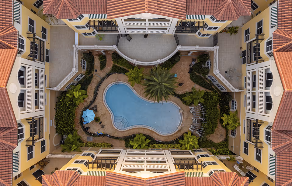 Aerial view of a central courtyard with a kidney-shaped swimming pool, palm trees, and surrounding yellow Mediterranean-style buildings with red tile roofs.