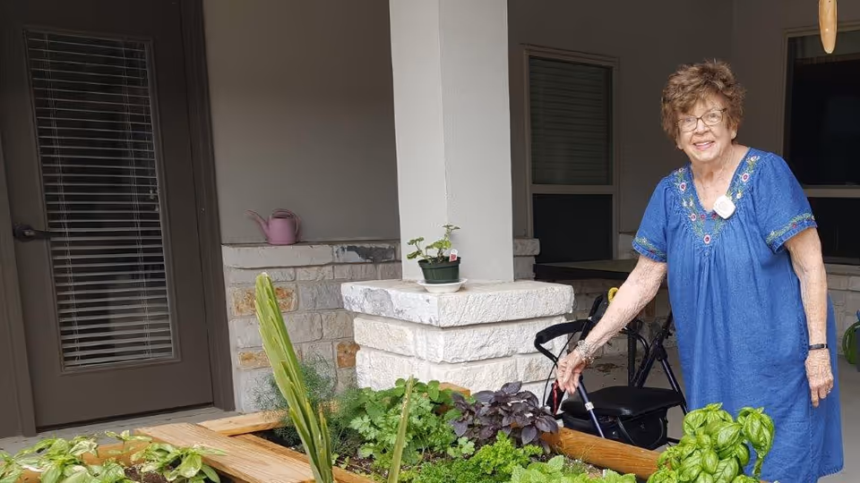 An elderly woman wearing a blue dress is standing next to a raised garden bed filled with various green plants and herbs. She is smiling and holding a gardening tool. The setting appears to be an outdoor patio area with stone pillars and a door with blinds in the background.