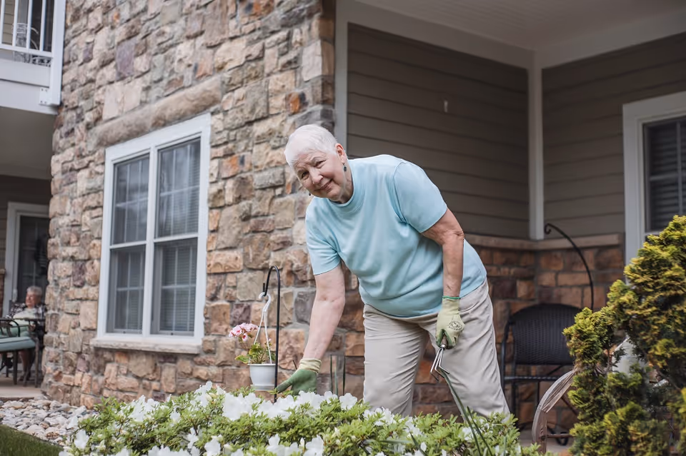 An elderly woman wearing gardening gloves tends to flowers outside a stone-faced senior living building.
