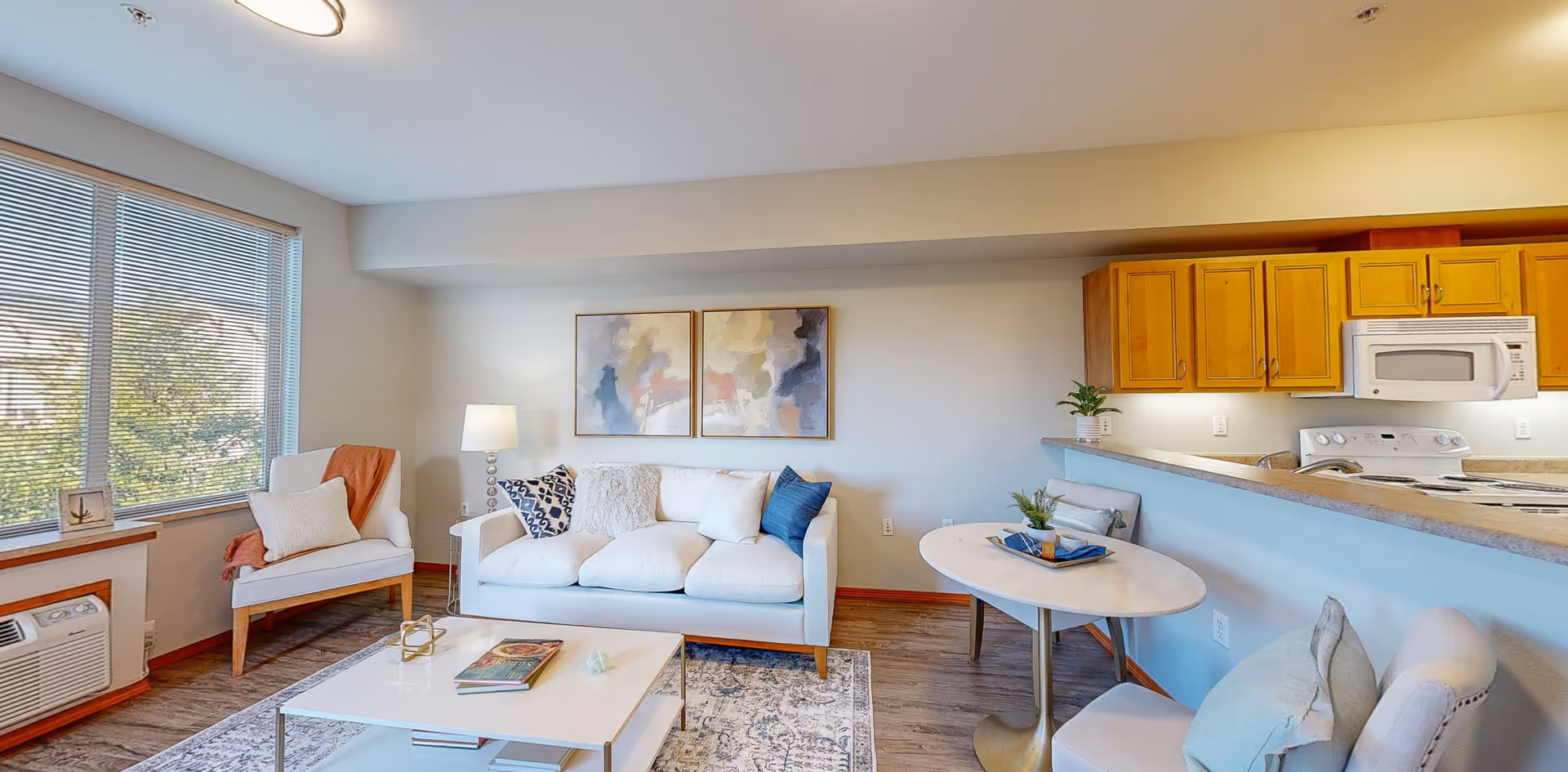 A bright living room and kitchen area in a senior living facility. The living room features a white sofa with multiple cushions, a white armchair with an orange throw blanket, a white coffee table with books, and a floor lamp. Two abstract paintings hang on the wall above the sofa. The kitchen has wooden cabinets, a white microwave, and a stove. A small round dining table with two chairs is positioned near the kitchen counter. Large windows with blinds allow natural light to fill the room.
