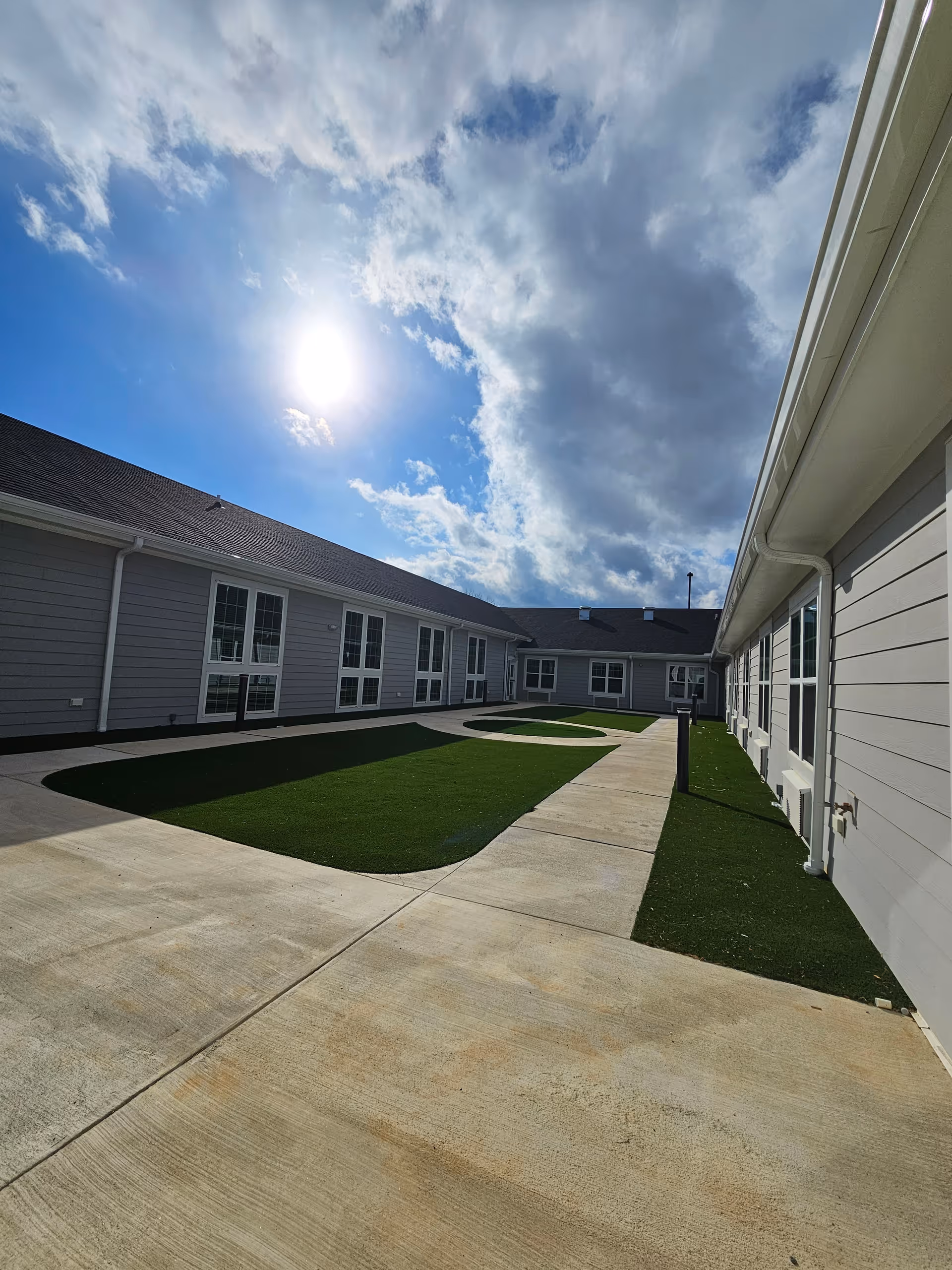 Outdoor courtyard area at Stonecroft facility with concrete walkways and green artificial turf patches, surrounded by single-story buildings with multiple windows under a partly cloudy sky with the sun shining brightly.