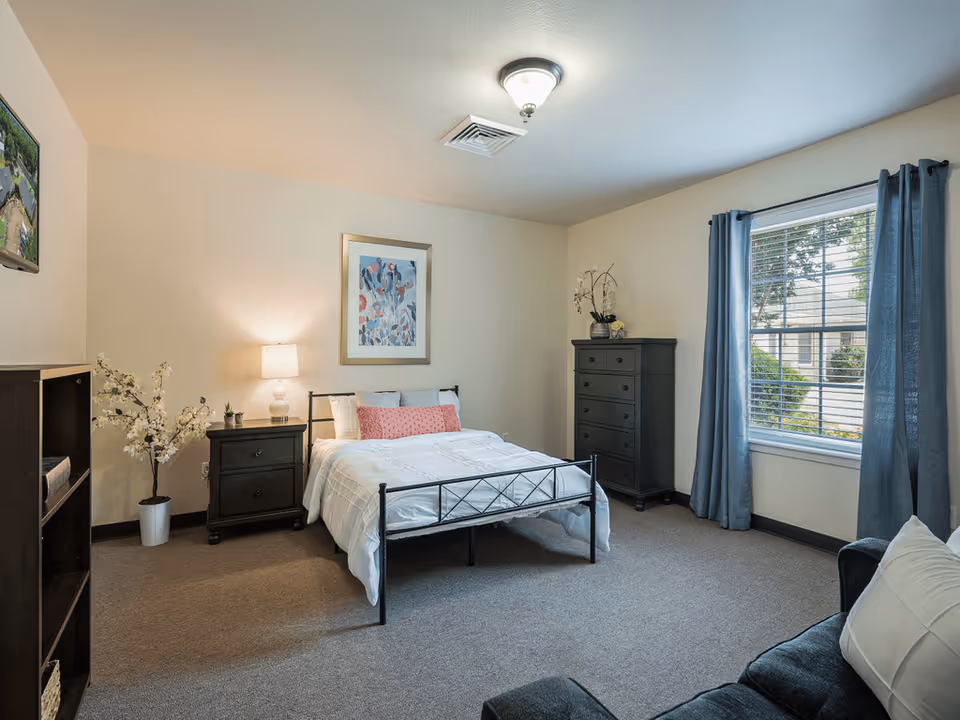 A cozy bedroom in an assisted living facility featuring a neatly made bed with white bedding and a pink accent pillow. The room includes a dark wooden nightstand with a lamp, a matching tall dresser with decorative plants on top, a window with blue curtains, a small bookshelf, and a comfortable chair with a white pillow. The walls are light-colored, and there is a framed floral artwork above the bed.
