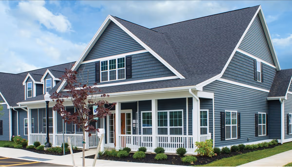 Exterior view of a two-story residential-style building with blue siding, white trim, multiple windows, and a covered porch with white railings. There is a small tree and landscaped bushes in front, along with a sidewalk and a streetlamp.