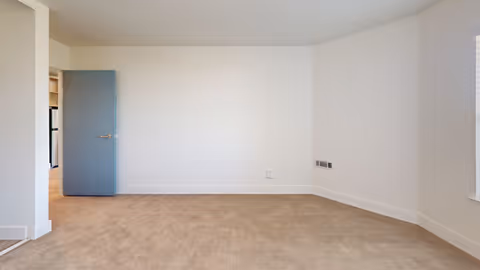 Empty room with beige carpet, white walls, a blue door slightly open leading to another room, and a window on the right side.
