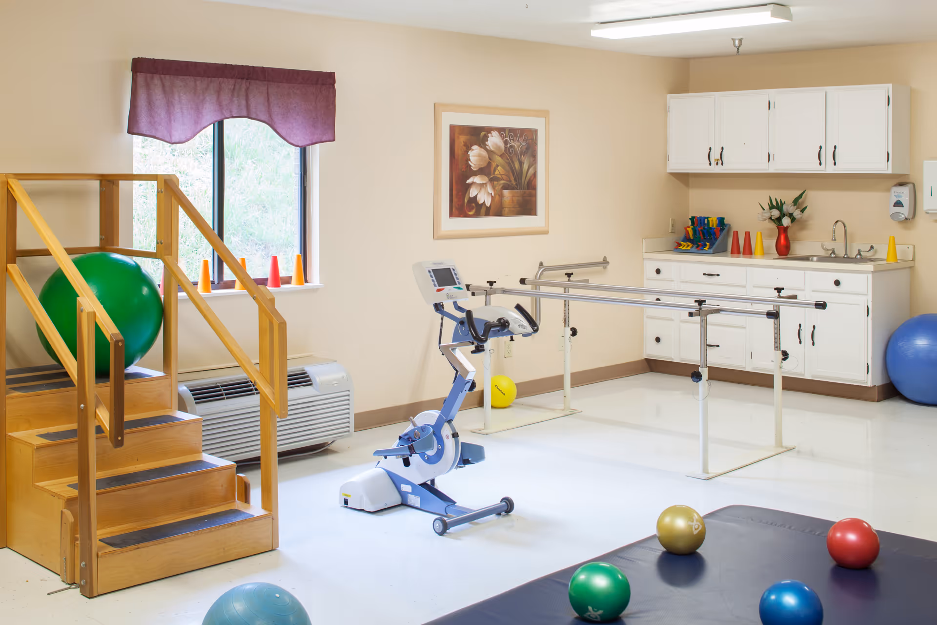 Bright rehabilitation room with parallel bars, an exercise bike, wooden therapy stairs, and colorful therapy balls.