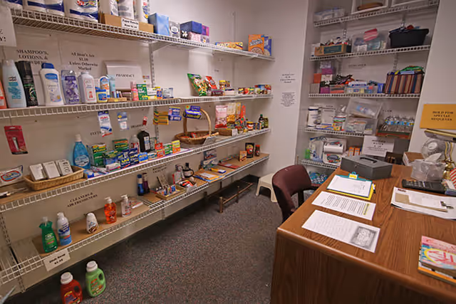 Small supply room with wire shelving stocked with toiletries, snacks, and household items and a desk with paperwork.