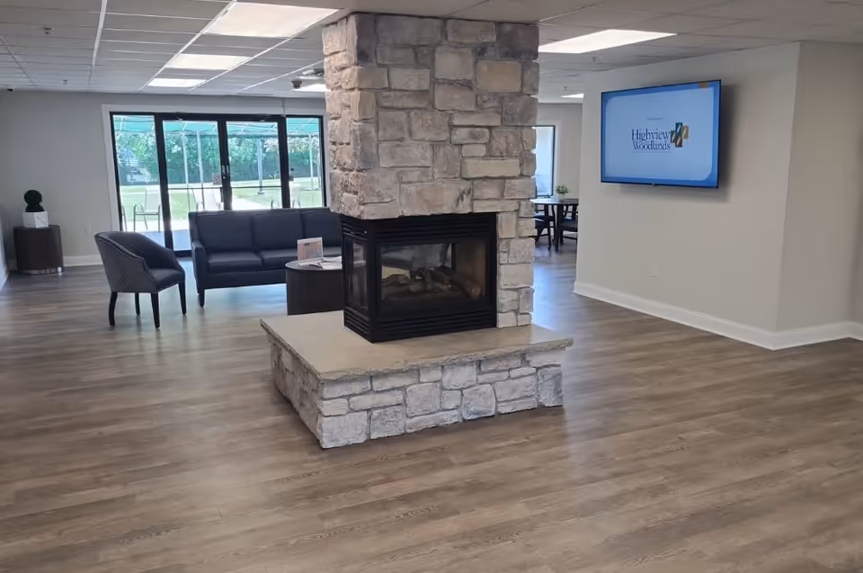 A spacious common area with a stone fireplace in the center, a dark blue sofa and a gray chair near large glass doors leading outside. A flat screen TV mounted on the wall displays the Highview In the Woodlands logo. The room has wood flooring and a bright, clean atmosphere.