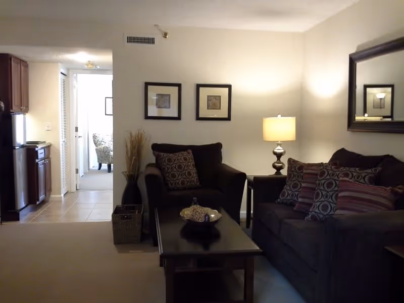 A cozy living room area with a dark armchair and sofa adorned with patterned cushions. A wooden coffee table with a decorative bowl sits in front of the seating. A table lamp on a side table provides warm lighting. Two framed pictures hang on the wall above the armchair, and a large mirror is mounted on the adjacent wall. In the background, a small kitchen area with wooden cabinets and a refrigerator is visible, along with a hallway leading to another room with an armchair near a window.